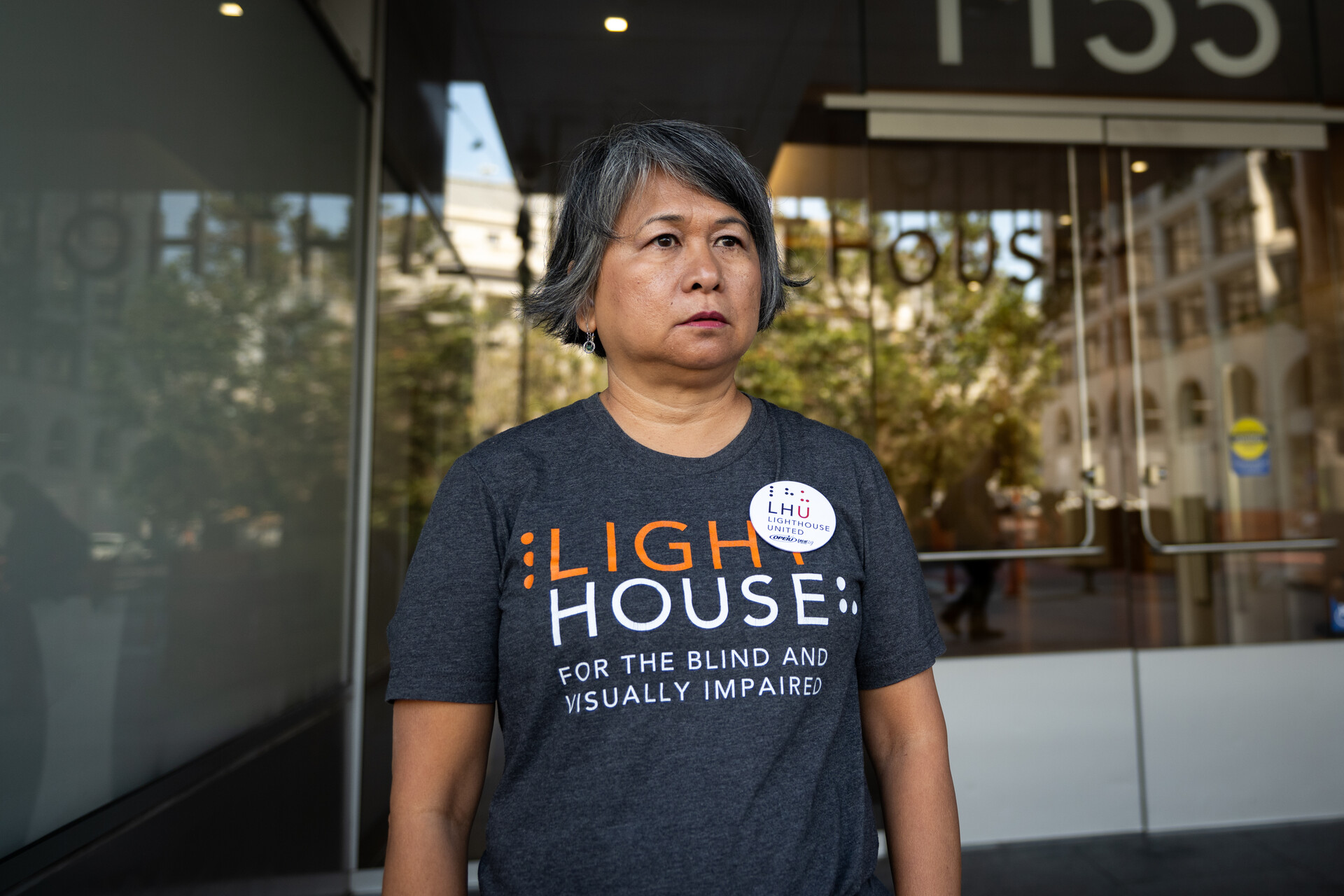 An Asian woman with short hair stands in front of a building wearing a LIGHTHOUSE shirt, looking to the side away from the camera.