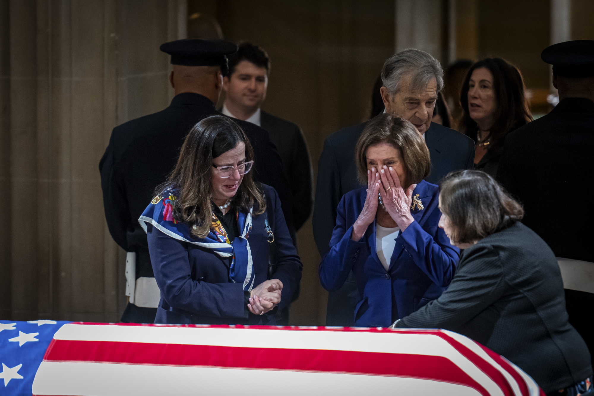 A person with long hair cries in front of a casket draped in an American flag.