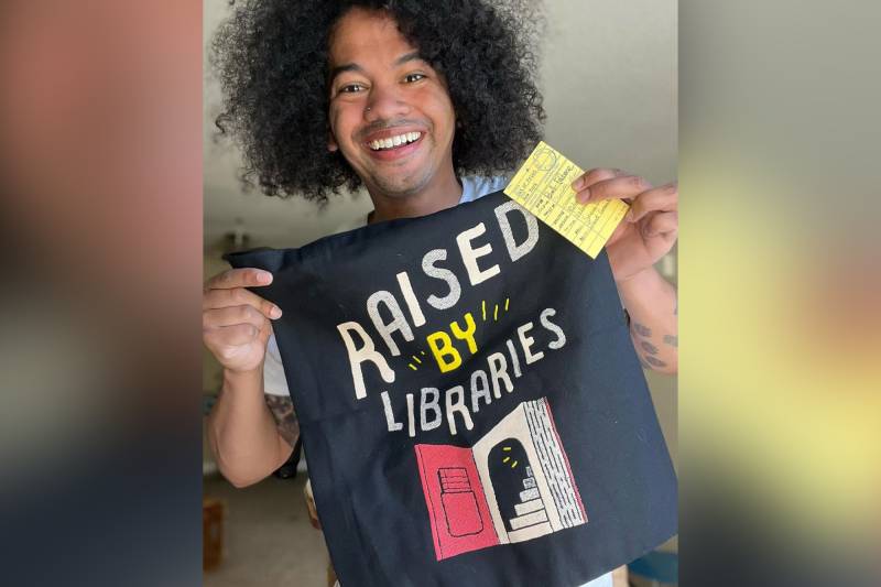 A person holds up a tote bag that reads "Raised by libraries" and smiles at the camera.