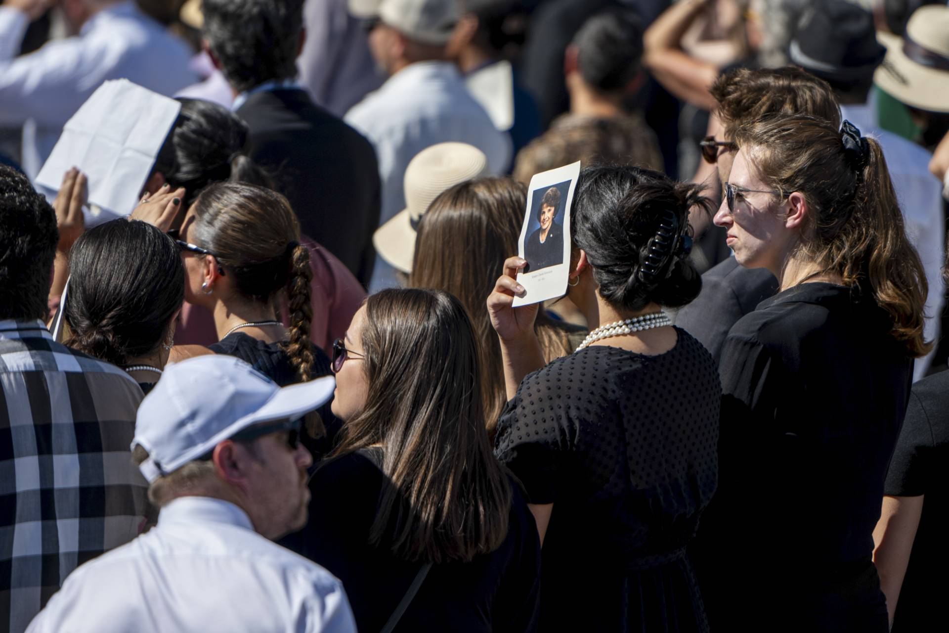 A large crowd of people and one person shielding their face from the sun using a piece of paper with the photo of a person on it.