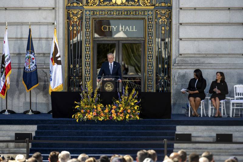 A person wearing a suit speaks at a podium in front of an ornate building with the words "City Hall" written above an entryway.