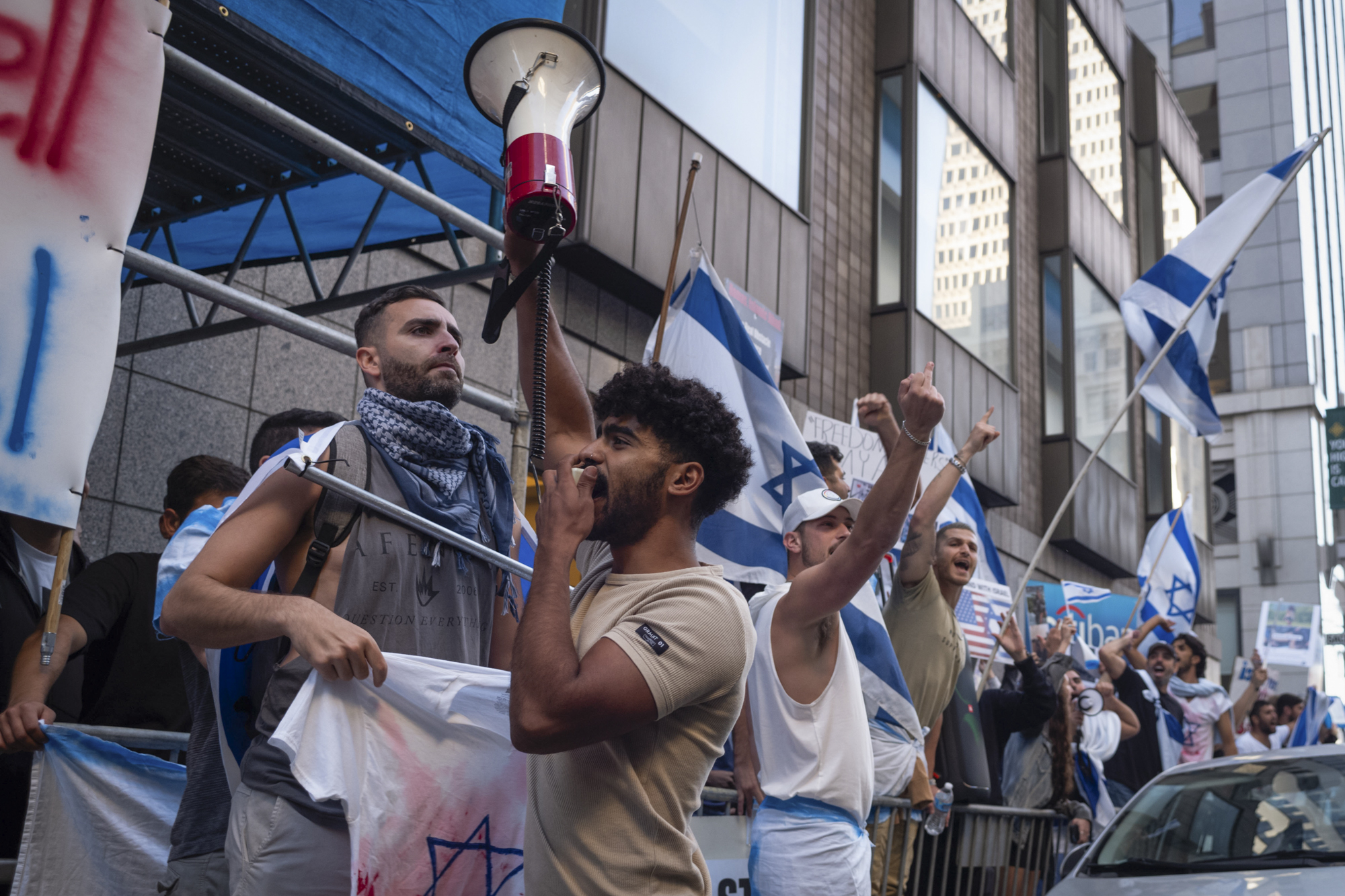 A group of people holding white and blue flags chant and yell in an urban setting.