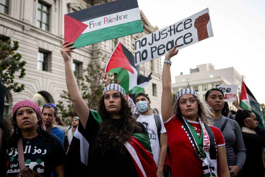 Two women wearing headscarves hold 'Free Palestine' and 'No justice, no peace' signs during a large outdoor rally.