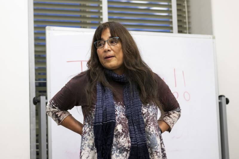 a Latina woman with long hair and glasses stands in front of a whiteboard