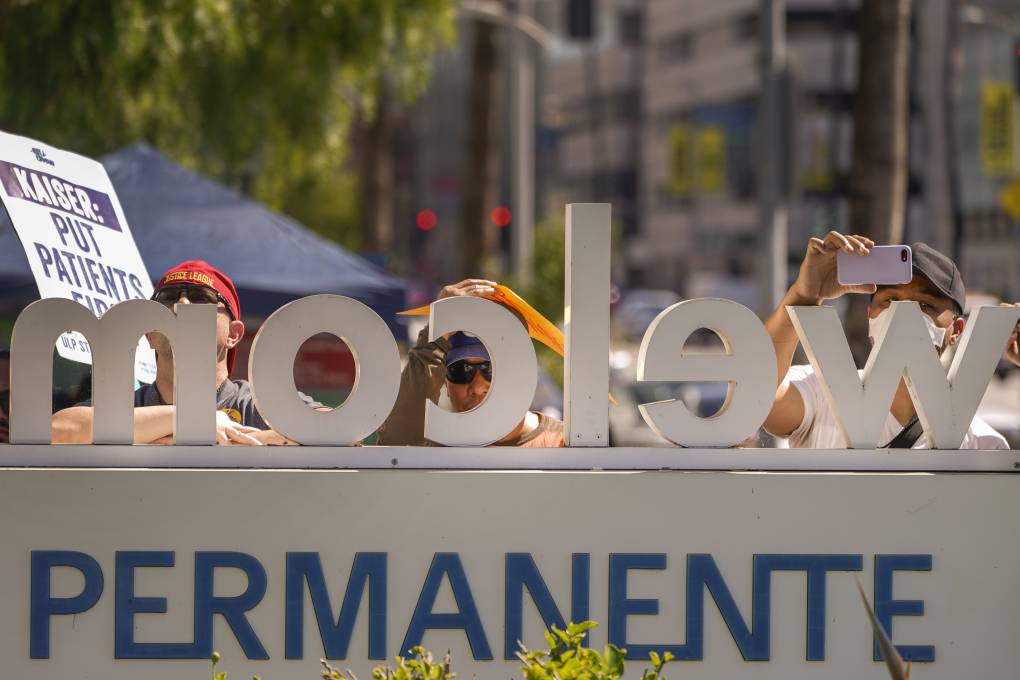 Protestors outside of a Kaiser Permanente in Los Angeles stand behind the medical center's sign holding signs.