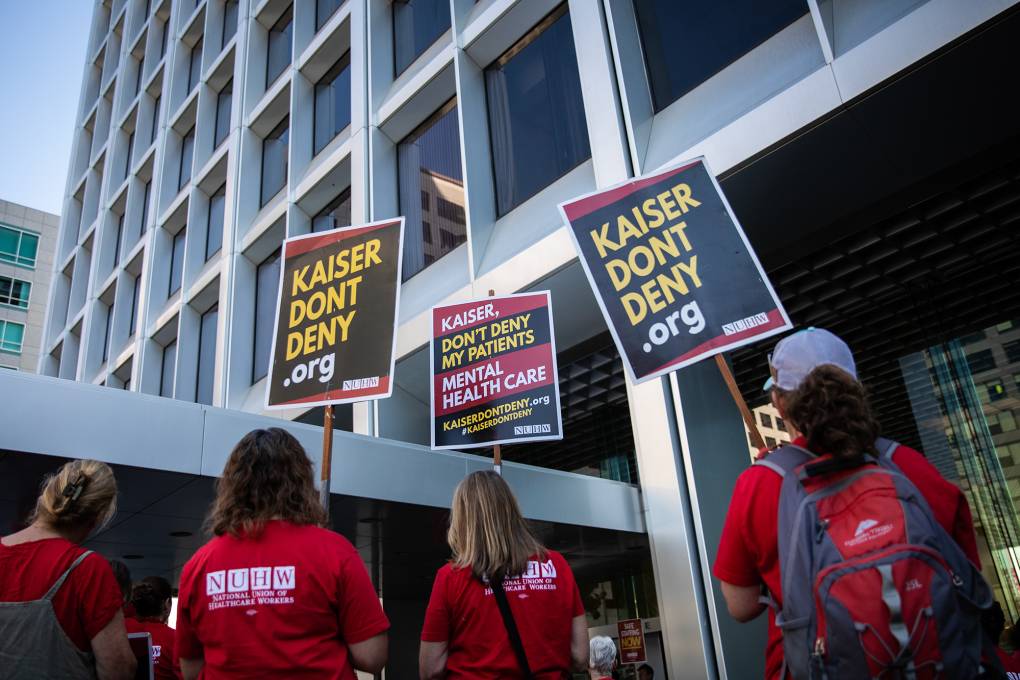 Mental health workers in red T-shirts hold signs in protest outside of a Kaiser facility in Oakland.