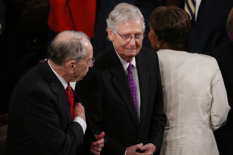 Two old white men wearing suits talk with each other on the floor of the US Senate