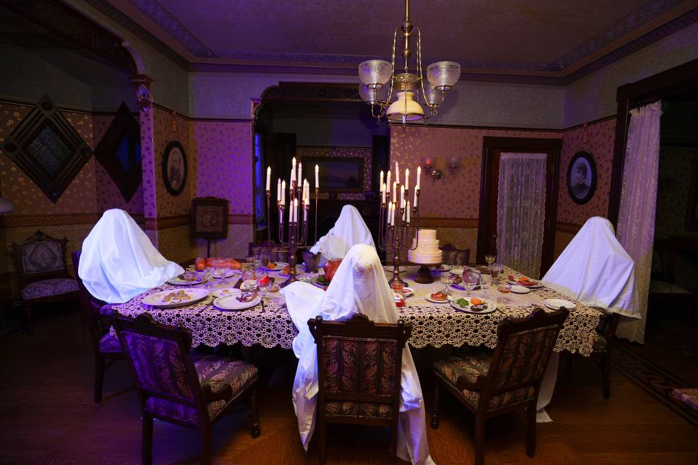 An ornately decorated Victorian dining room with a table in the center. Around the table are seating 4 fake ghosts, covered in white sheets. There are two large candelabras on the table as well as a fake dinner.