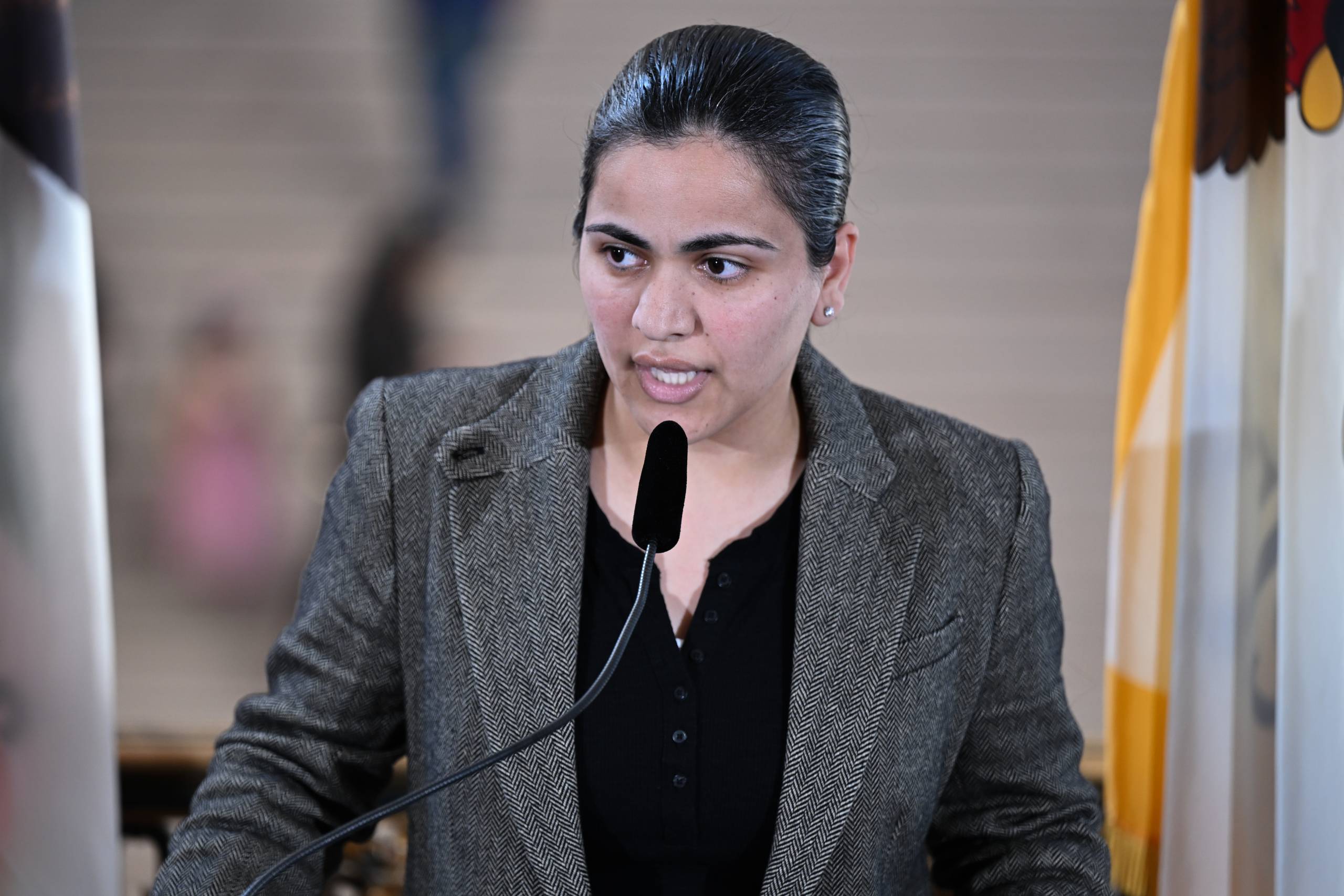 A South Asian woman in a gray suit speaks into a mic.