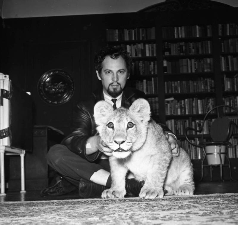 Black and white image of a man with dark curly hair and a goatee seated cross legged on the floor of a room. In front of him is a lion cub, which looks at the camera.