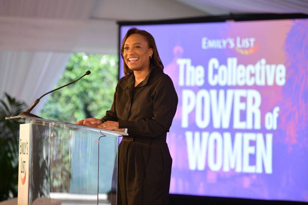 An African American woman dressed in a black suit standing at a podium with a sign that reads "Emily's List The Collective Power of Women" in the background.