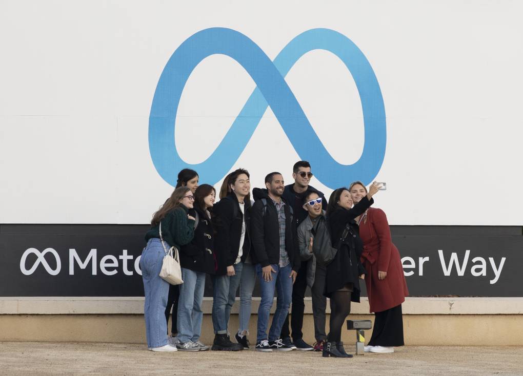 A group of young people stand in front of Meta headquarters and take a selfie in front of the company logo, which is akin to an infinity symbol.