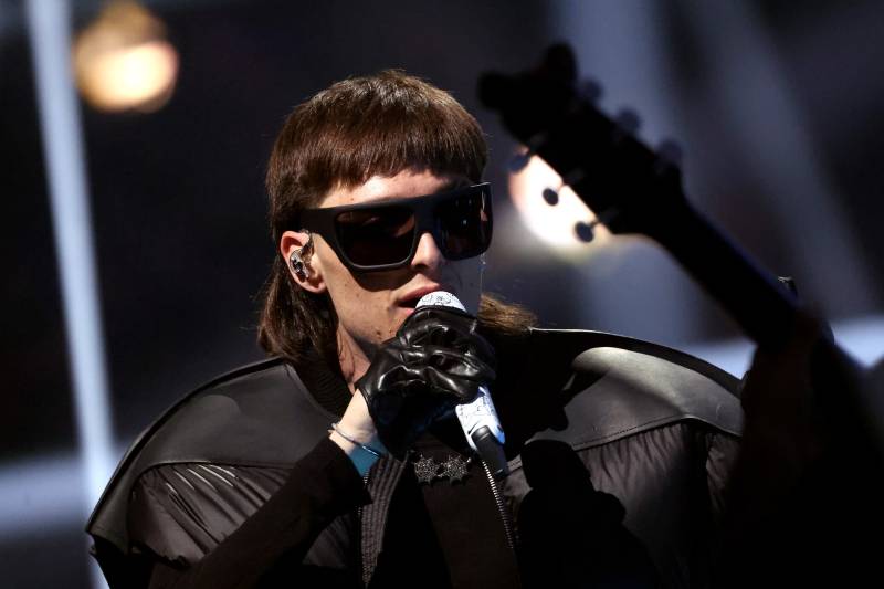 A younger man with blunt brown bangs and mullet wears large dark sunglasses and a broad-shouldered black shirt, singing into a mic onstage