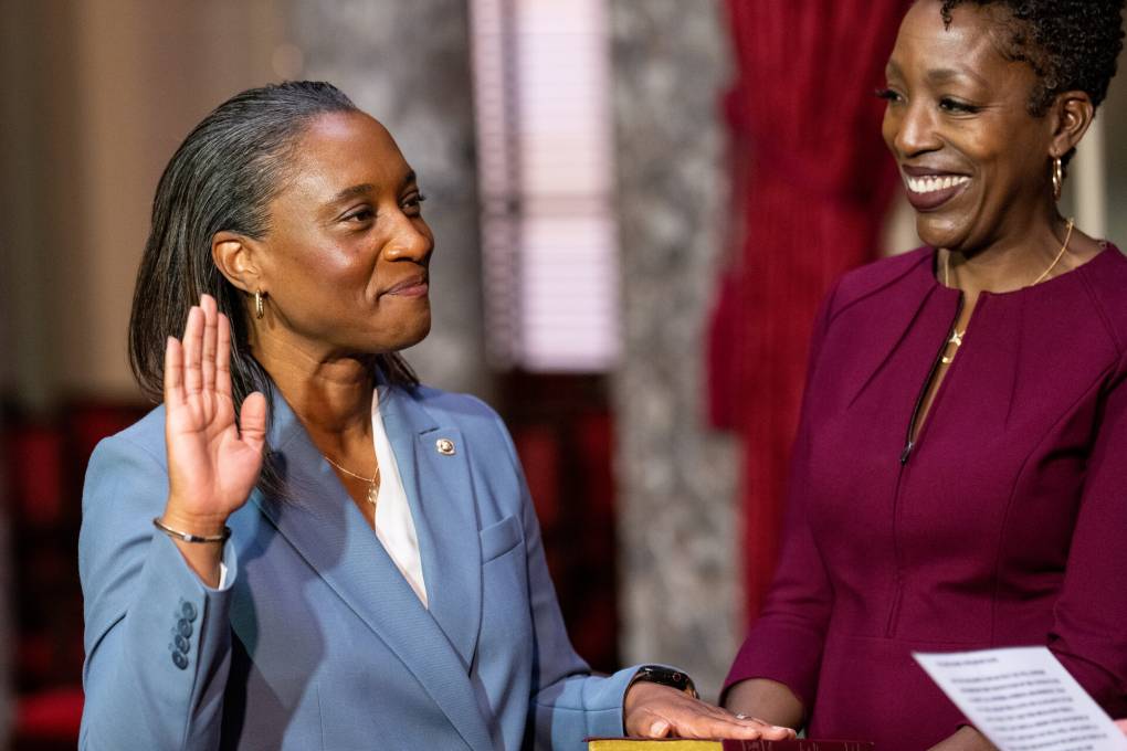 a smiling Black woman wearing a light blue blazer smiles with her right rand raised as another smiling Black woman wearing a maroon blouse looks on