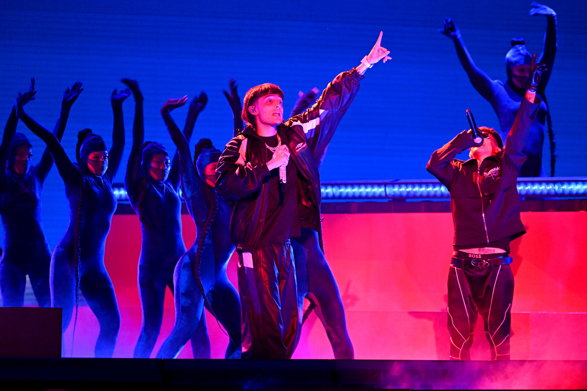 A group of people perform on a blue and red toned stage, with red lighting cast on their faces and bodies, holding their arms in the air. Two men at the front hold microphones.