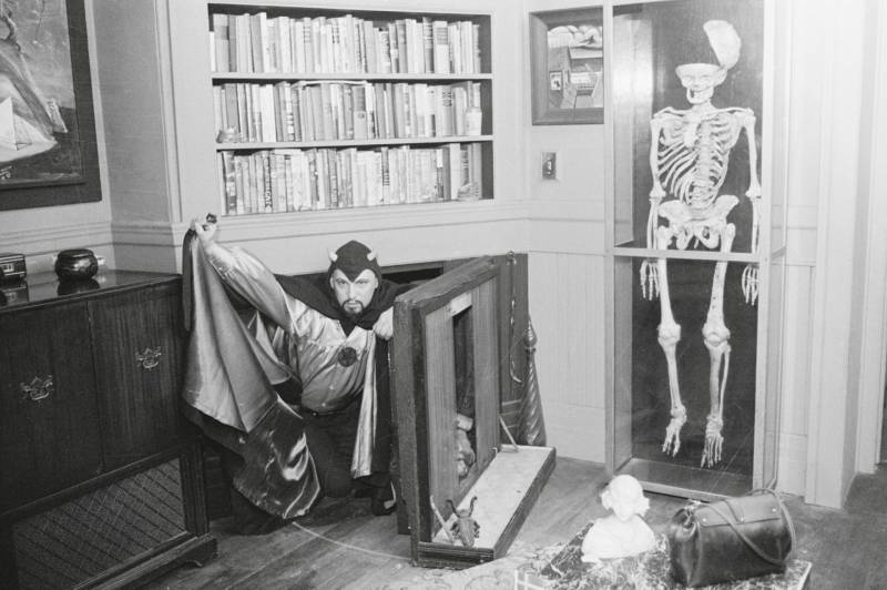 A mean in a hooded devil costume and cloak emerges from a secret passage way in the fireplace of a a room in his house. There are bookshelves above hi, and a human skeleton mounted in a large display case on the wall to the right.