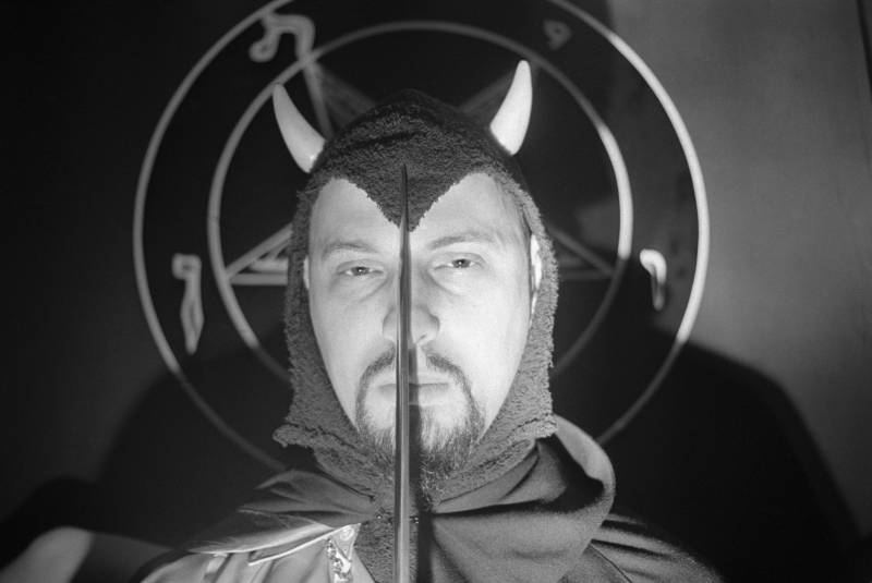 Black and white close up image of a man wearing a hooded cloak with devil horns. He is holding the point of a sword in front of his face. He casts a shadow onto the wall behind him, which features a large pentagram.