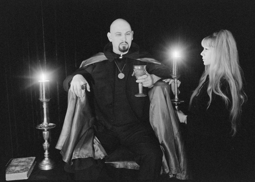A black-and-white image of a man with a polished bald head wearing all black and a large cloak stands center, holding a chalice in one hand. To his right, a woman with long blonde hair holds a candle aloft. The pair are illuminated by candlelight in an otherwise dark room.