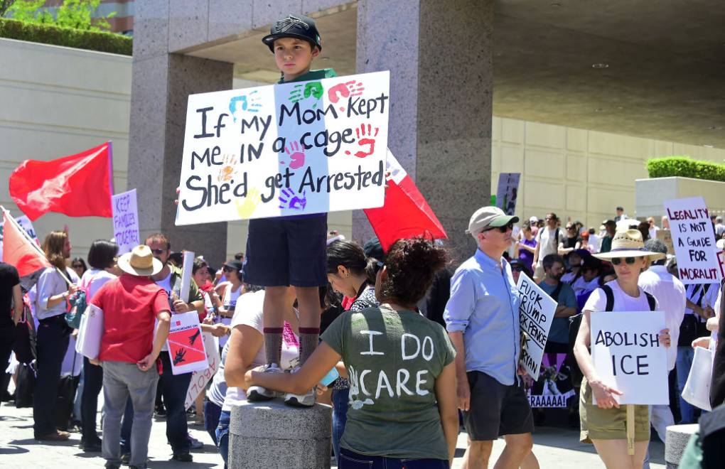 A young boy stands on top of a small barrier in a crowd of people with a sign that reads "If my Mom kept me in a cage, she'd get arrested."
