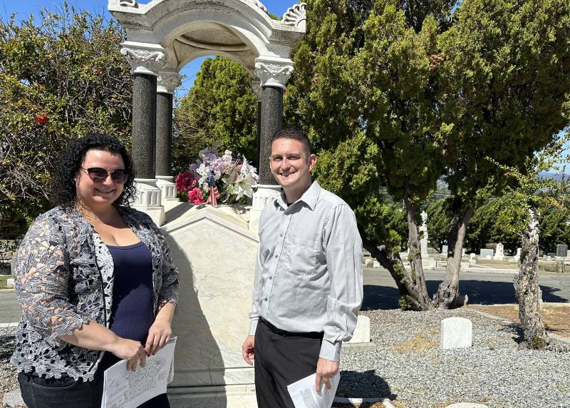Caroline Sandoval and Gary Winni stand next to a tall white grave marker in a cemetery. Flowers rest on top.