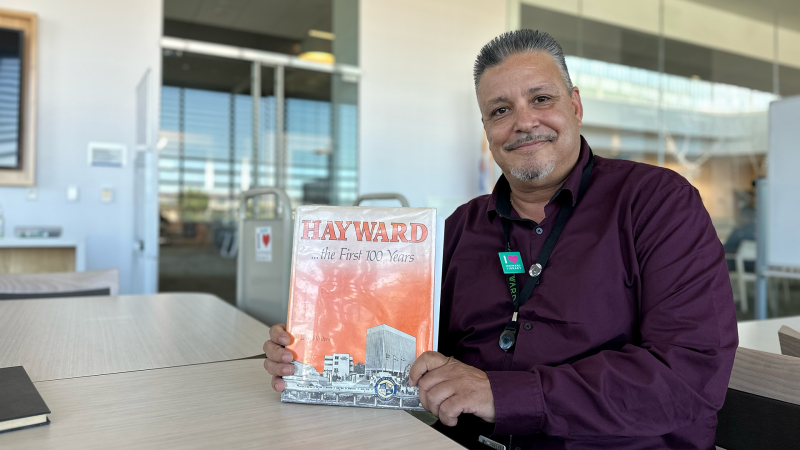 Hector Villasenor sits smiling at a table in the library holding a book called "Hayward... the first 100 years."