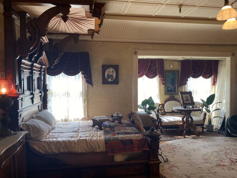 A preserved Victorian era bedroom with an ornately carved wooden headboard, sitting area, and lace curtains.