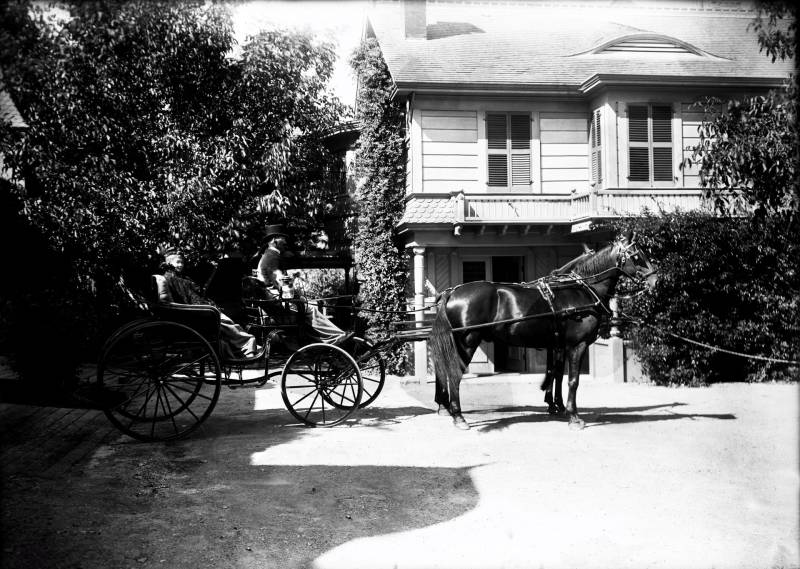Black and white archival image of a woman sitting in the back of a horse drawn carriage. The man at the front of the carriage is wearing a top hat, and it is pulled by 2 horses. It sits unmoving in front of a house, with trees to either side of the image.