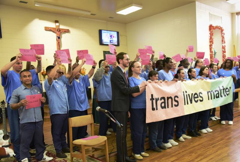 A group of people, most wearing the same uniform of blue shirts and pants, stand together holding signs and joined by a person wearing a suit helping to hold a banner reading "trans lives matter."