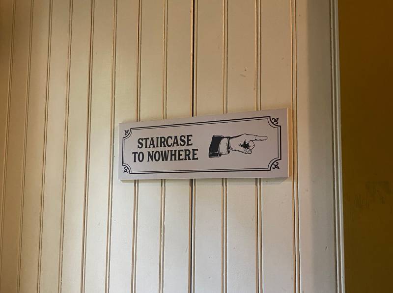 A sign on a cream colored wood paneled wall that says "Staircase to nowhere." The sign also includes an illustration of a hand pointing to the right.
