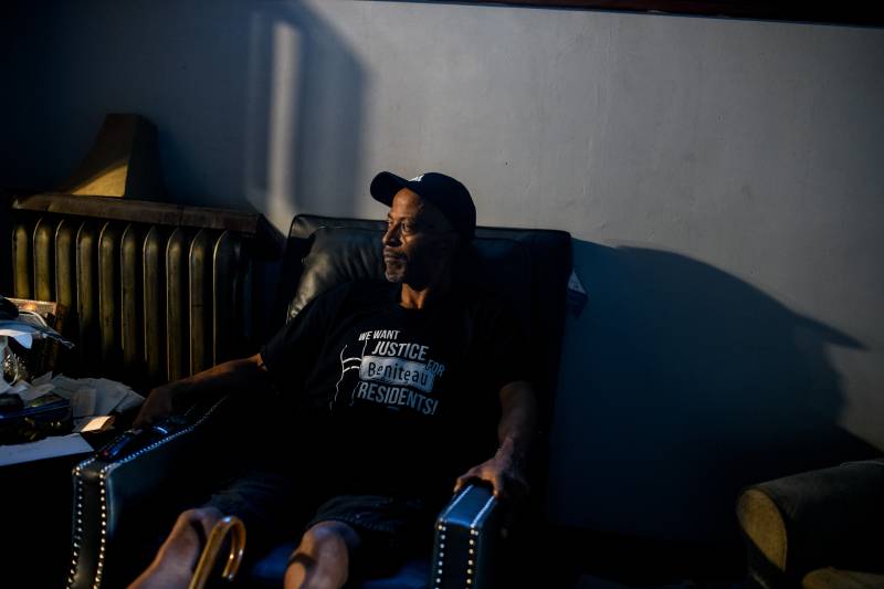 A Black man wearing a cap and t-shirt sits in a chair looking to the side.