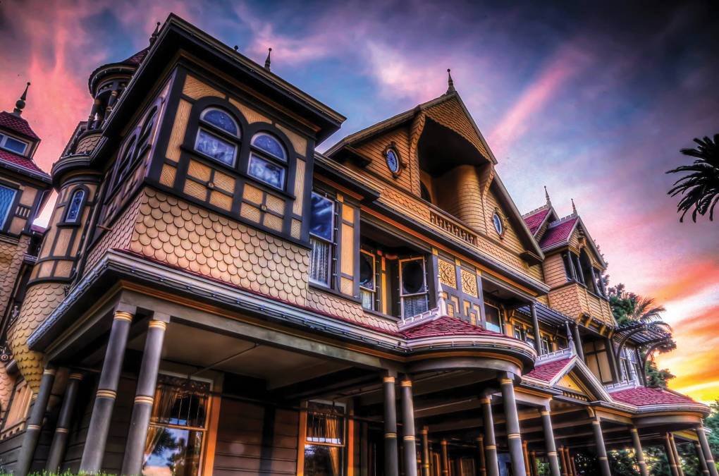 Looking up at an elaborate Victorian mansion with yellow, black and purple detailing. The sky behind the house is lit with the colors of a sunset.
