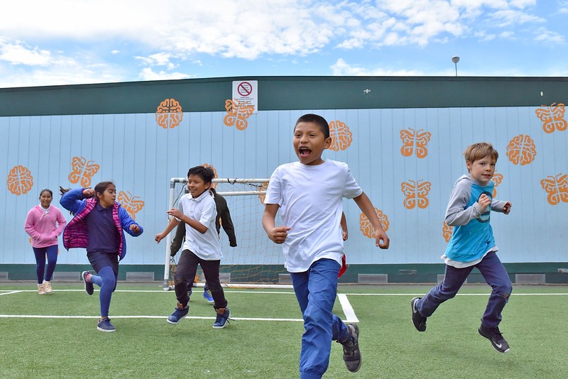 Children are running around at recess on the schoolgrounds. A building with painted orange butterflies is seen in the background.