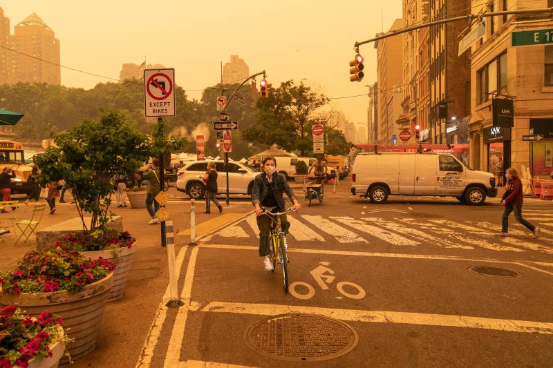 A man wearing a face mask rides a bike in a city with cars and people moving in the background.
