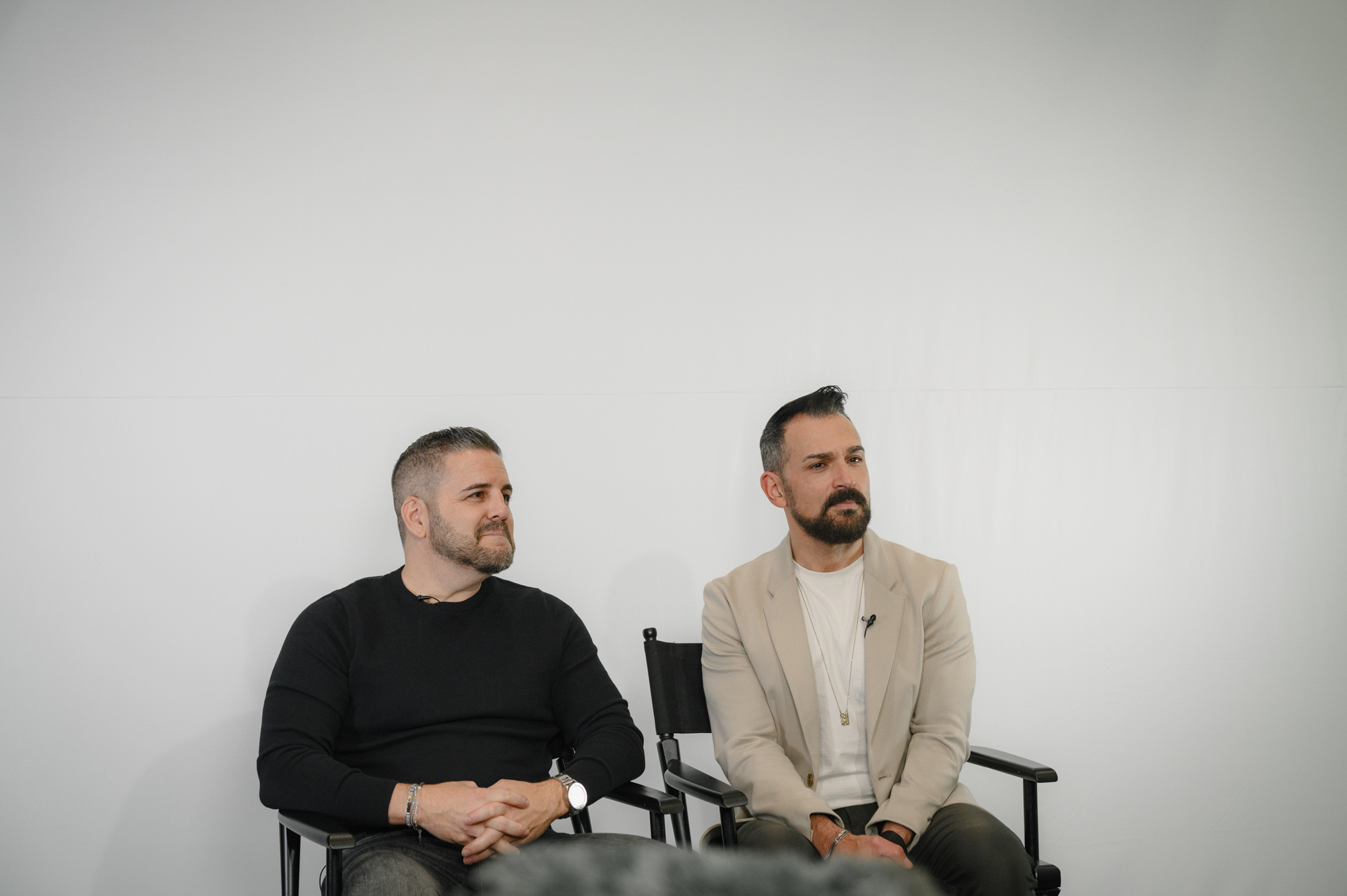 Two people sit next to each other in chairs in front of a white backdrop.