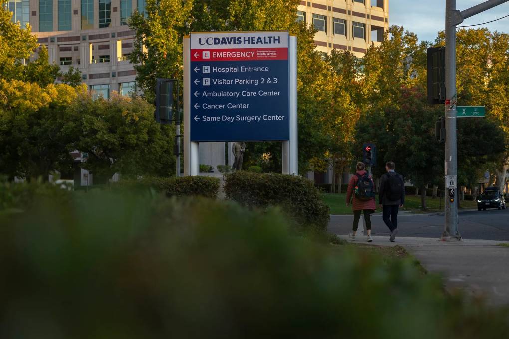 Two people walk past a sign that says 'UC Davis Health' - with a large hospital in the background.