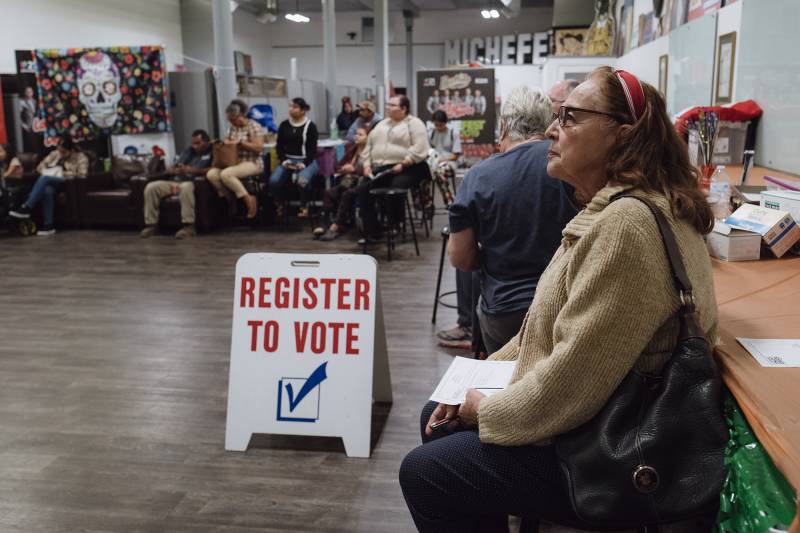 A side view of a woman wearing glasses seated next to other people with a sign that reads "Register to Vote" on the floor.
