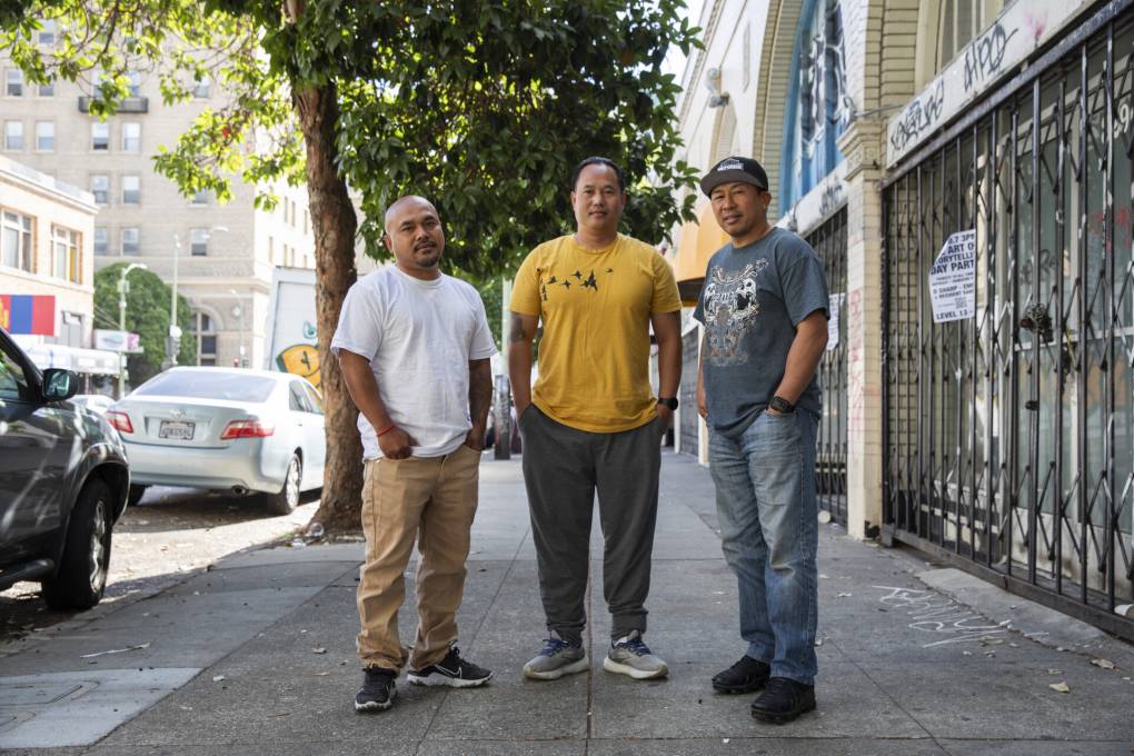 A photo portrait of three middle-aged Southeast Asian men in T-shirts standing on a sidewalk.