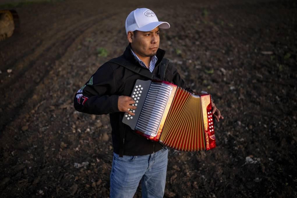 A man in a baseball cap stands in fallow fields and plays the accordion.