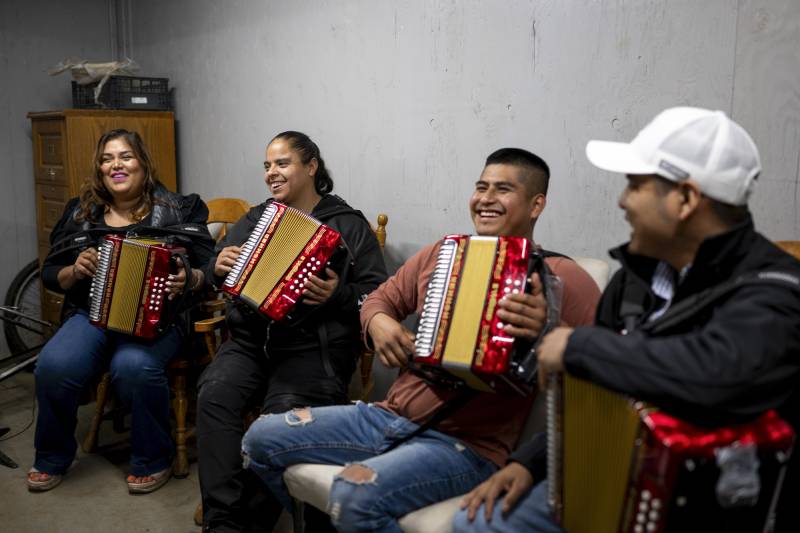 A group of people smile while sitting together indoors and holding bright red accordions.