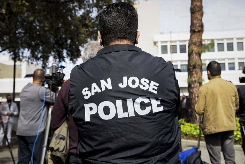 A person wears a black San José Police jacket in an outdoor setting.