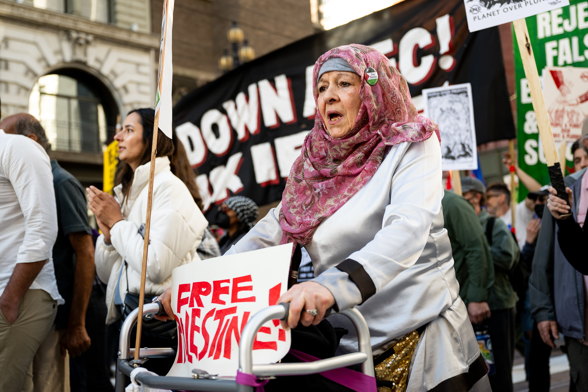 An older woman uses a walker with a sign that says "free Palestine" in a large crowd.