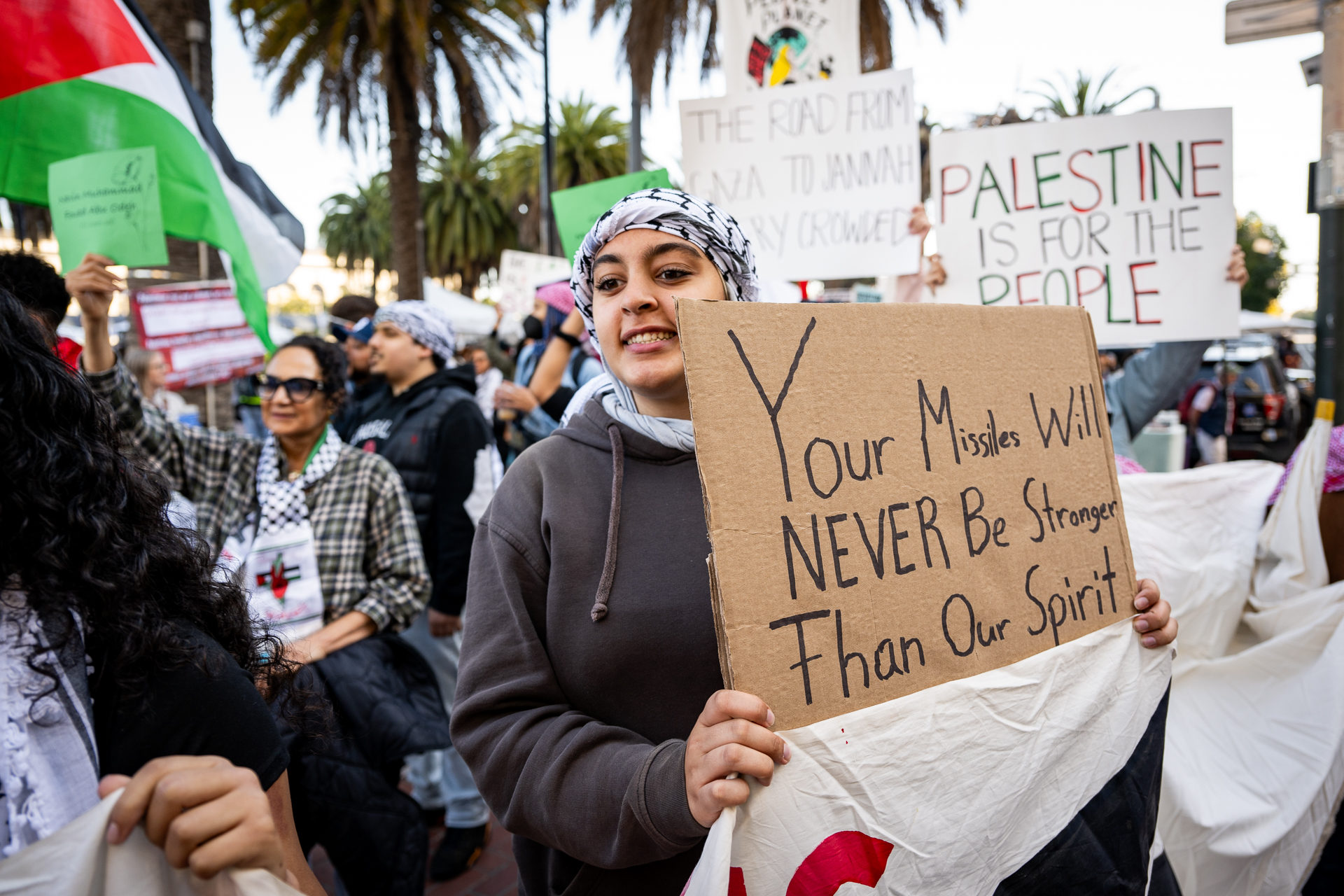 A young woman holds a sign that says "your missiles will never be stronger than our spirit" in a crowd of people.