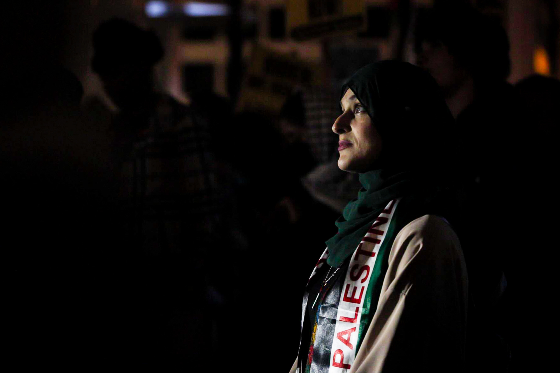 A woman is highlighted by a light at night wearing a Palestine scarf and hijab.