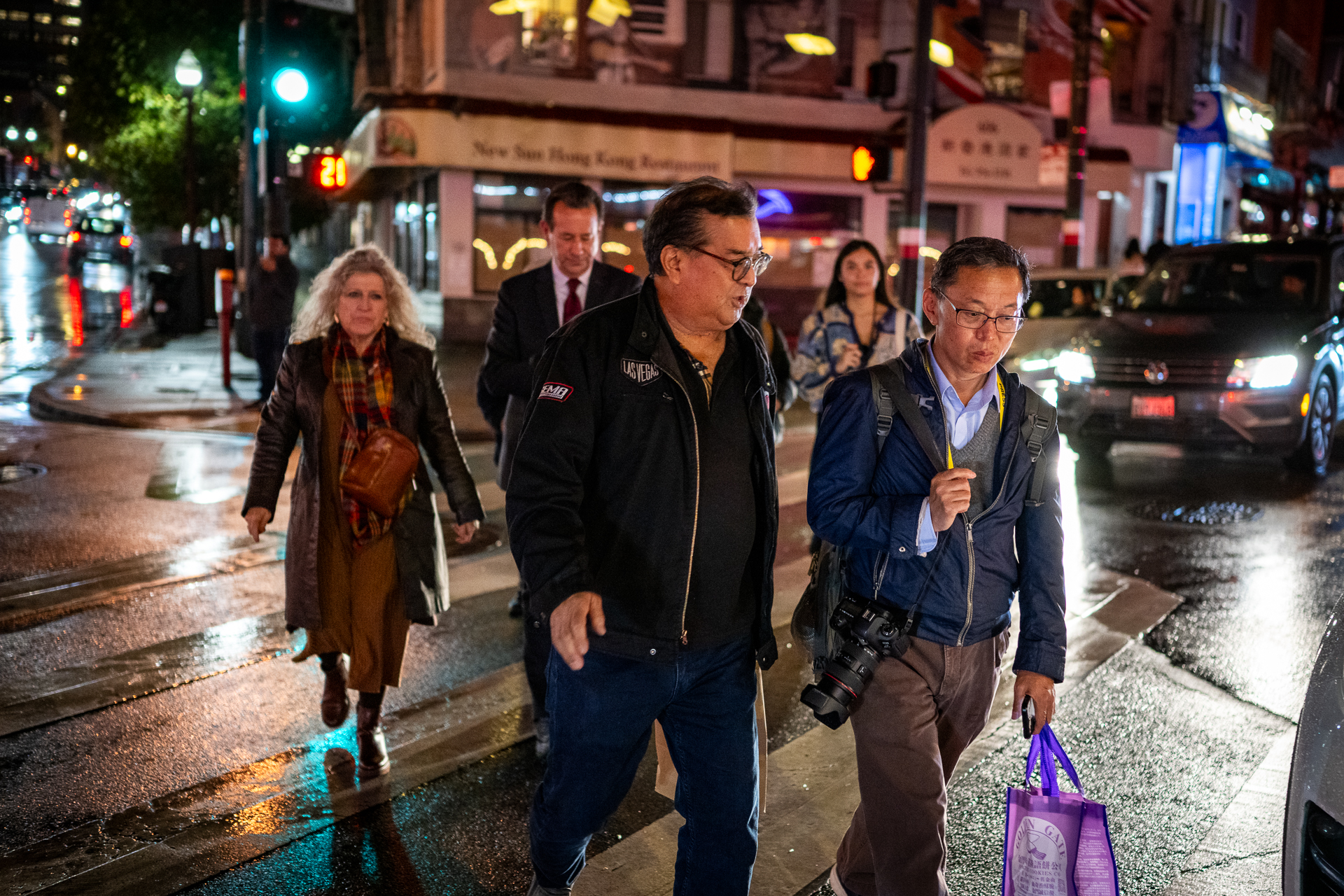 A group of people walk on a crosswalk across a wet city street.