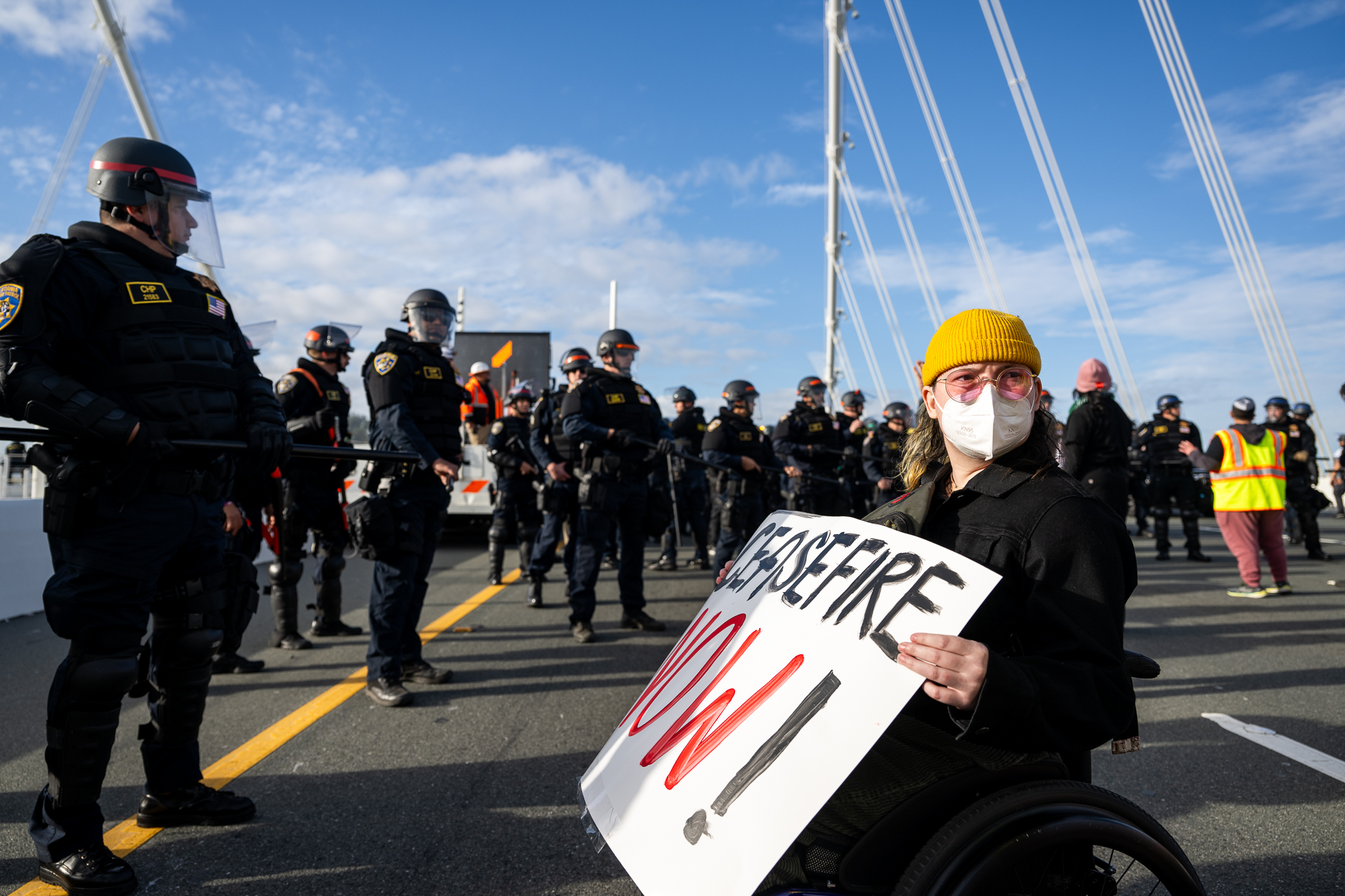 A person in a wheelchair holds a protest sign with a line of police on the left and behind them.