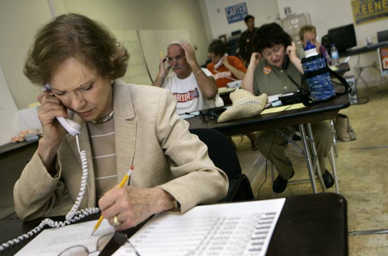 A vintage photo of a woman wearing a tan blazer holding a phone to her ear with a pencil in the other hand over a piece of paper at a desk with others seated behind her.