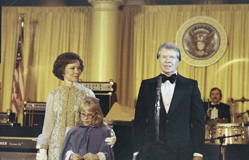 A man in a tuxedo, a woman in a light colored dress and a young woman in a purple dress stand.