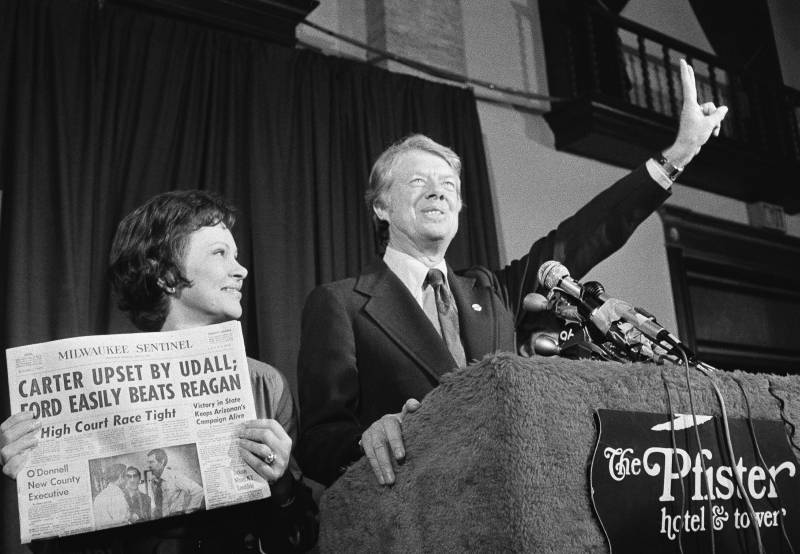A black and white photo of a woman holding up a newspaper next to a man in a suit with one hand in the air at a podium.