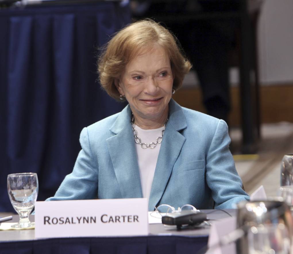 An older woman in a light blue blazer jacket with a sign that says "Rosalynn Carter."