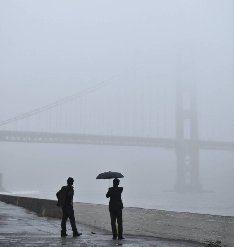 Two people stand silhouetted on a cement walkway while the Golden Gate Bridge is just barely visible through thick fog in the background.
